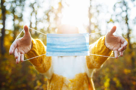 Young woman wearing an orange hat and sweater holds the medical sterile mask in her hands in the autumn forest. Female breathes fresh air in fall park during coronavirus covid19 pandemic. Healthcareの写真素材