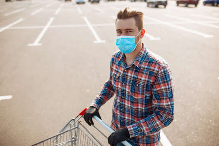 Young man stands outside the supermarket with the trolley wearing sterile medical mask and gloves to protect from coronavirus. Customer at a shops parking is going for purchases. Healthcare conceptの写真素材