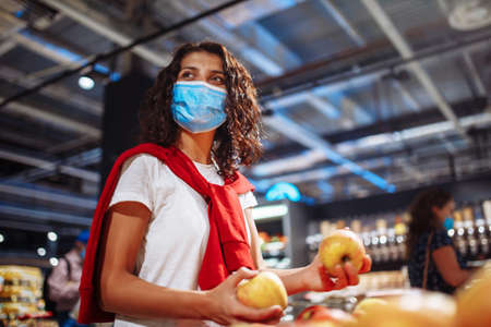 Young woman picks up some vegitables and fruits at a supermarket wearing a medical sterile mask...