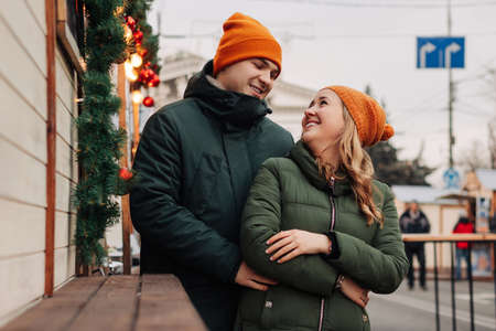 Young happy couple having fun together at a Christmas market. Attractive man and woman wearing orange hats and green jackets hug and kiss at the street at New Year eve. Winter mood conceptの写真素材