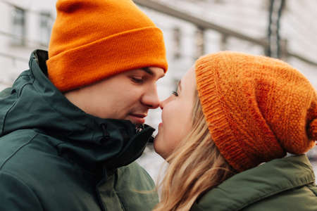 Young happy couple having fun together at a Christmas market. Attractive man and woman wearing orange hats and green jackets hug and kiss at the street at New Year eve. Winter mood conceptの写真素材