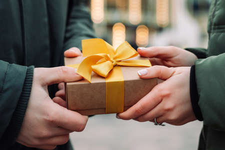 Hands of a couple with a gift present. Man gives a small present with a yellow ribbon to his girlfriend on the street. Woman takes a gift from her boyfriend. Valentines day and Christmas conceptの写真素材