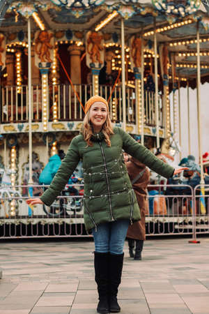Young woman's portrait at a Christmas market decorated with lights. Female wearing orange hat and green coat stands at a seasonal New Year market. Winter mood conceptの写真素材