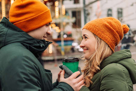Young beautiful couple having fun together outdoors holding coffee and feeling warm and happy during winter holidays. Christmas lights and garlands at the market. Love and festival conceptの写真素材