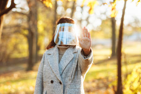 Young woman wearing a face shield to protect from coronavirus desease stands in the park during autumn season on a sunset. Female outdoors shows no sign with hand. Quarantine pandemic fall conceptの写真素材