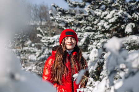 Portrait of a girl teenager wearing red coat standing among fir trees covered with snow at the winter park. Young girl having fun in the winter forest. Winter vibes concept.の写真素材