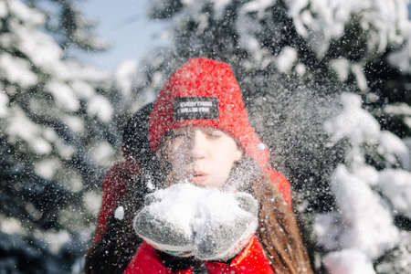 Close portrait of a girl teenager wearing red jacket and hat and blowing the snow out of her hands at the winter park. Young woman having fun on a bright winter day in the forest. Happiness concept.の写真素材