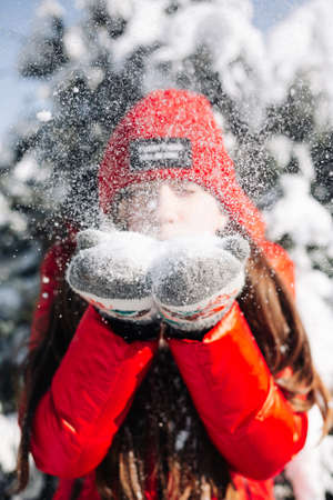 Close portrait of a girl teenager wearing red jacket and hat and blowing the snow out of her hands at the winter park. Young woman having fun on a bright winter day in the forest. Happiness concept.の写真素材