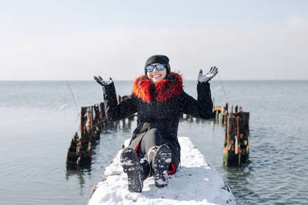 Portrait of a girl wearing black and red coat sits and having fun on a snowy pier at sea during frosty cold winter day. Young woman smiling and throwing snow in the air. Happiness concept.の写真素材