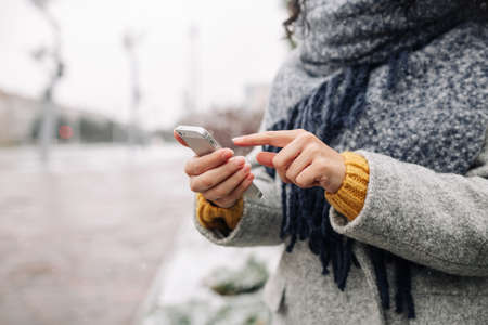 Young woman using her mobile phone at a snowy winter park. Closeup of female checking news and...