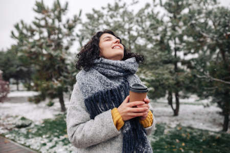 Young woman wearing frey fashion coat and blue scarf stands with a coffee to go at a winter snowy park. Female enjoys hot drink during frosty cold day outdoors. Joy and holidays conceptの写真素材