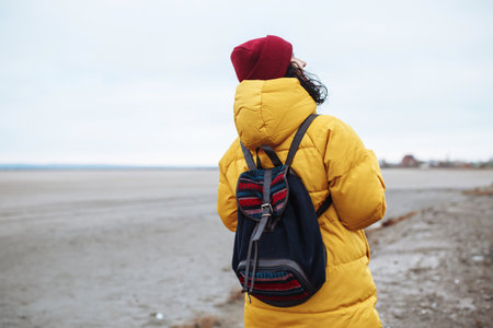 Backview of a young woman tourist with a backpack walking on sideroad among vast empty winter valley lowland. Female traveler wearing yellow jacket and red hat. Hitch-hiking, traveling conceptの写真素材