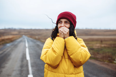 Beautiful girl wearing red hat and yellow jacket warms her hands with her breath on the lonely road....