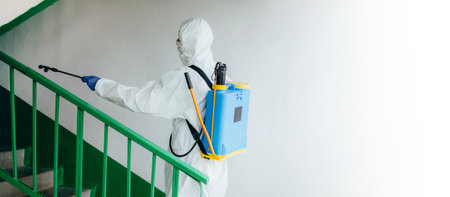 Wide shot. Sanitary professional worker in protective suit disinfects a staircase entryway block of flats. Coronavirus prevention measures at residential areas. New normal and stop Covid-19 spread.の写真素材