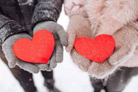 Red paper hearts in the hands of a loving couple outside at a snowy winter park. Romantic man and woman celebrate Valentine's day with symbols of love. Sign of 14 Februaryの写真素材