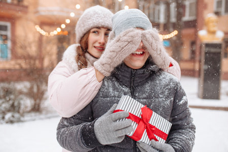 Young romantic woman closes boyfriend's eyes with her hands in woolen mittens to make a surprise outside at a winter snowy park. Smiling man holds a present box with red bow. Valentine's day conceptの写真素材