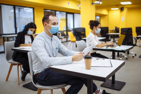 Three colleagues work together at an office keeping social distance weaing medical masks. People sit at the desks working hards to support business. New normal, covid-19 pandemic measures conceptの写真素材