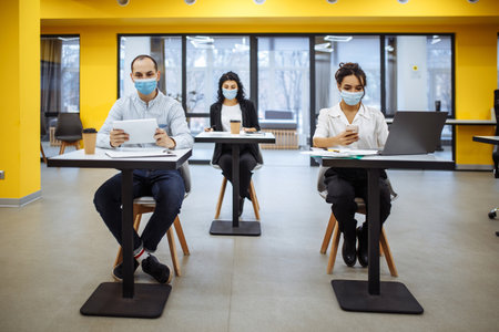 Three colleagues work together at an office keeping social distance weaing medical masks. People sit at the desks working hards to support business. New normal, covid-19 pandemic measures conceptの写真素材