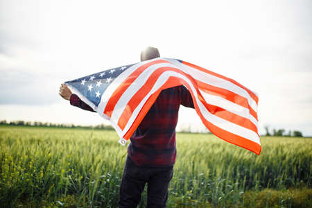 Young man waves an american flag at the green field of wheat. Male celebrates USA independence day on a sunny day. Holidays conceptの写真素材