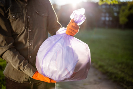 Young man volunteer hold a trash bag in his hands outdoors wearing gloves. A volunteer cleans up the park on a sunny bright day. Clearing, pollution, ecology and plastic conceptの写真素材