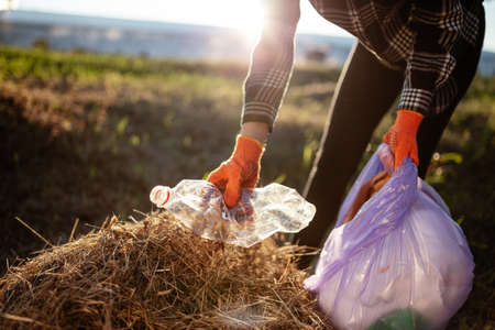 Woman wearing gloves collects used plastic bottle and throws it into trash bag. A volunteer cleans up the park on a sunny bright day. Clearing, pollution, ecology and plastic conceptの写真素材