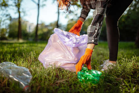 A woman collects and puts used plastic bottle into a trash bag. A volunteer cleans up the park on a...