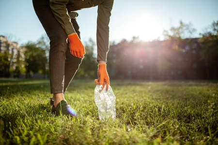 Young man volunteer collects used plastic bottle from the ground outdoors wearing gloves. Volunteer...
