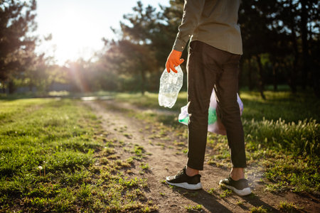 Young man walks with trahs bag and used plastic bottle in his hands. A volunteer cleans up the park on a sunny bright day. Clearing, pollution, ecology and plastic conceptの写真素材
