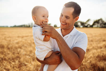 A father holds his little son on the hands in the middle of the golden wheat field. Man with his kid having fun outside on the bright sunny day. Family values and happiness conceptの写真素材