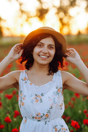Portrait of a smiling girl in a poppy field. Happy woman in white dress and hat on a sunset background. Sunny photo of a brunette in a ripe fieldの写真素材