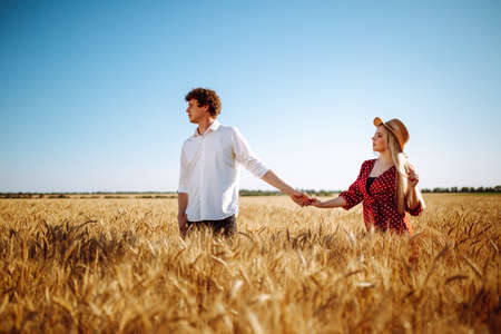 Happy man and woman are walking at sunset in a wheat field. A loving couple holding hands at sunset. A young girl in a dress with polka dots and a guy in a white shirtの写真素材