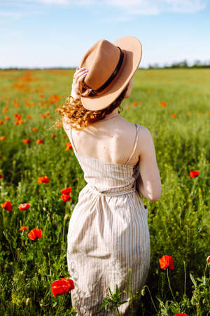 Girl posing in a poppy field. A woman in a hat with beautiful long hair stands in the middle of a beautiful poppy garden. Girl in a poppy meadow with blooming flowers. Spring landscapeの写真素材
