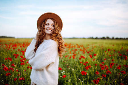 Beautiful girl in a white sweater in a poppy field. Happy woman in hat posing in flower field. A girl with beautiful curly hair in the middle of a field with blooming poppiesの写真素材