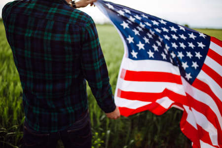 Independence Day. A man in a plaid shirt with an American flag in a field. Patriotic young man in a wheat field. 4th of July. Freedom and independence concept. Holidays, patriotism, pride, freedomの写真素材