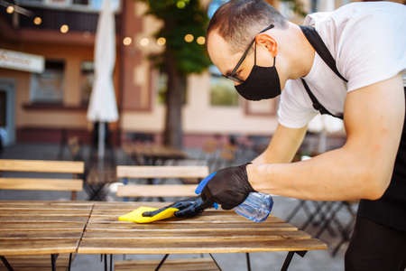 Side view portrait of waiter in protective mask and gloves wiping wooden table with yellow rag at outdoor cafe. New normal concept.の写真素材