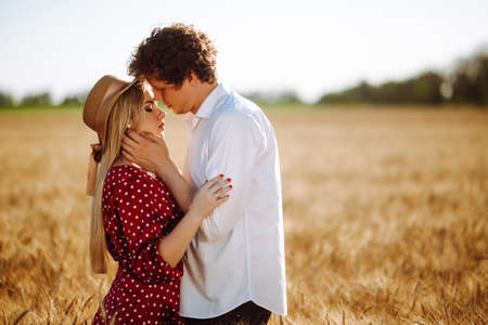 Portrait of a couple in love against the backdrop of the setting sun. A girl and a guy in wheat field. A man in white shirt and a woman in a dress with polka dots pose in the middle of a summer fieldの写真素材