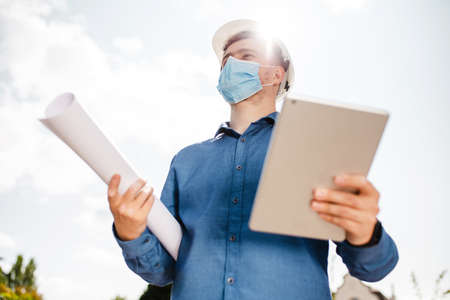 Young architect man wearing contractor helmet and protective mask holding project paper plan and...