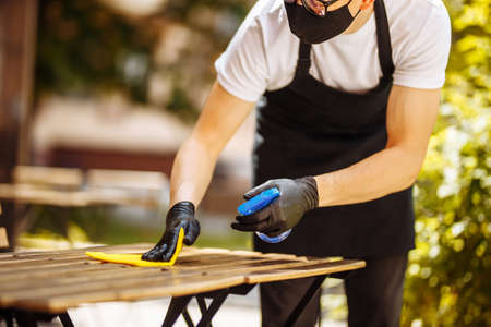 A waiter in a protective mask and gloves clean table with disinfectant and wipe before welcome customer. Pandemic health riskの写真素材