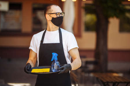 Young restaurant worker wearing black medical mask holds a tray with rug and spray with sanitizer on it. Waiter at a cafe prepares for cleaning before visitors and clients comeの写真素材