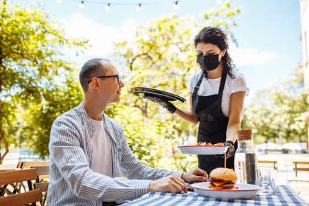 A young waitress wearing a protective mask and gloves brought the order to a visitor. Cafe terrace,...
