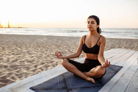 Young caucasian girl doing yoga asana in the beach with eyes closed. Young woman in lotus position in sportswear against the backdrop of the sunset and sea. Happy woman practicing yoga on the beachの写真素材
