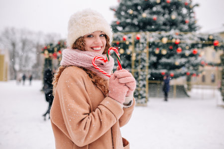 A beautiful curly girl with Christmas candies in her hands poses against the background of a New Year tree on a beautiful snowy winter dayの写真素材
