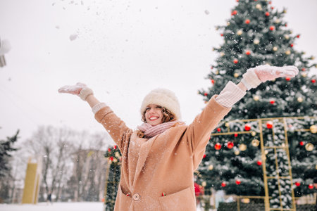 A curly girl blows snow against the backdrop of a beautiful New Year tree on a beautiful winter day. A woman plays in the snow in Decemberの写真素材