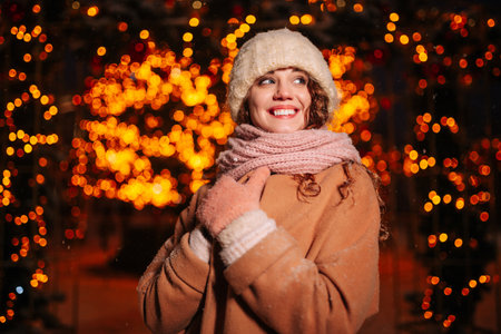 Portrait of a young curly girl in a knitted hat and scarf posing against the background of Christmas lights and on a pleasant winter eveningの写真素材