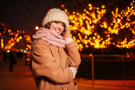 Portrait of a young curly girl in a knitted hat and scarf posing against the background of Christmas lights and on a pleasant winter eveningの写真素材