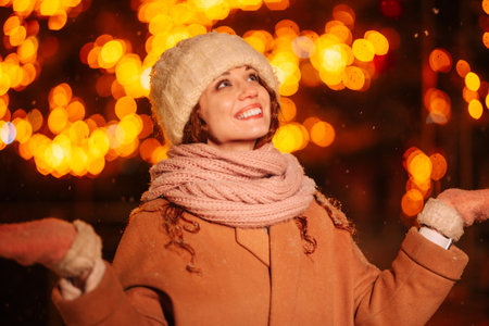 A captivating curly-haired young lady in the evening, framed by the glow of lights and a decorated Christmas tree. A nighttime stroll through the festive city on Christmas Eveの写真素材