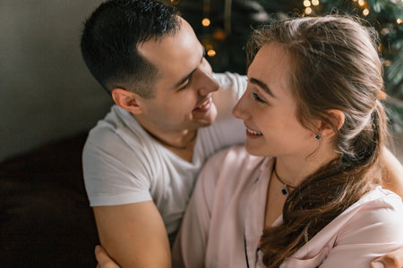 A young and beautiful couple hugging each other against the backdrop of a Christmas tree. The man and woman, dressed in pajamas, joyfully spend the New Year holidays at homeの写真素材