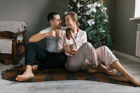 A young and happy couple sits on a cozy warm blanket near the Christmas tree at home. The beautiful man and woman enjoy hot chocolate and spend the New Year holidays in a comfortable home atmosphereの写真素材