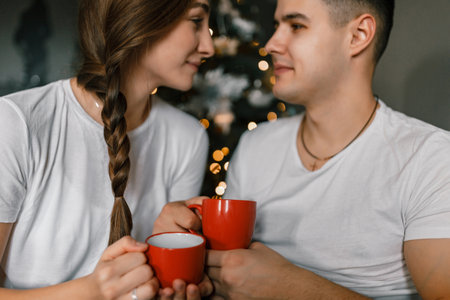 A young and happy couple sits near the Christmas tree at home. The beautiful man and woman enjoy hot chocolate and spend the New Year holidays in a comfortable home atmosphereの写真素材