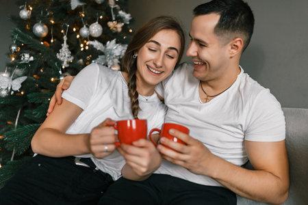 A young and happy couple sits near the Christmas tree at home. The beautiful man and woman enjoy hot chocolate and spend the New Year holidays in a comfortable home atmosphereの写真素材
